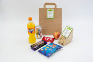 Small brown paper bag containing a business lunch of sandwich, drink, crisps, chocolate bar and an apple